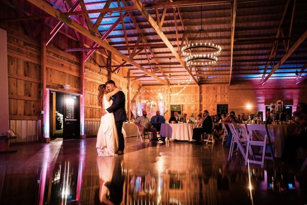 Couple sharing their first dance at a wedding reception in a rustic barn setting, with wooden walls, elegant lighting, and seated guests enjoying the celebration.