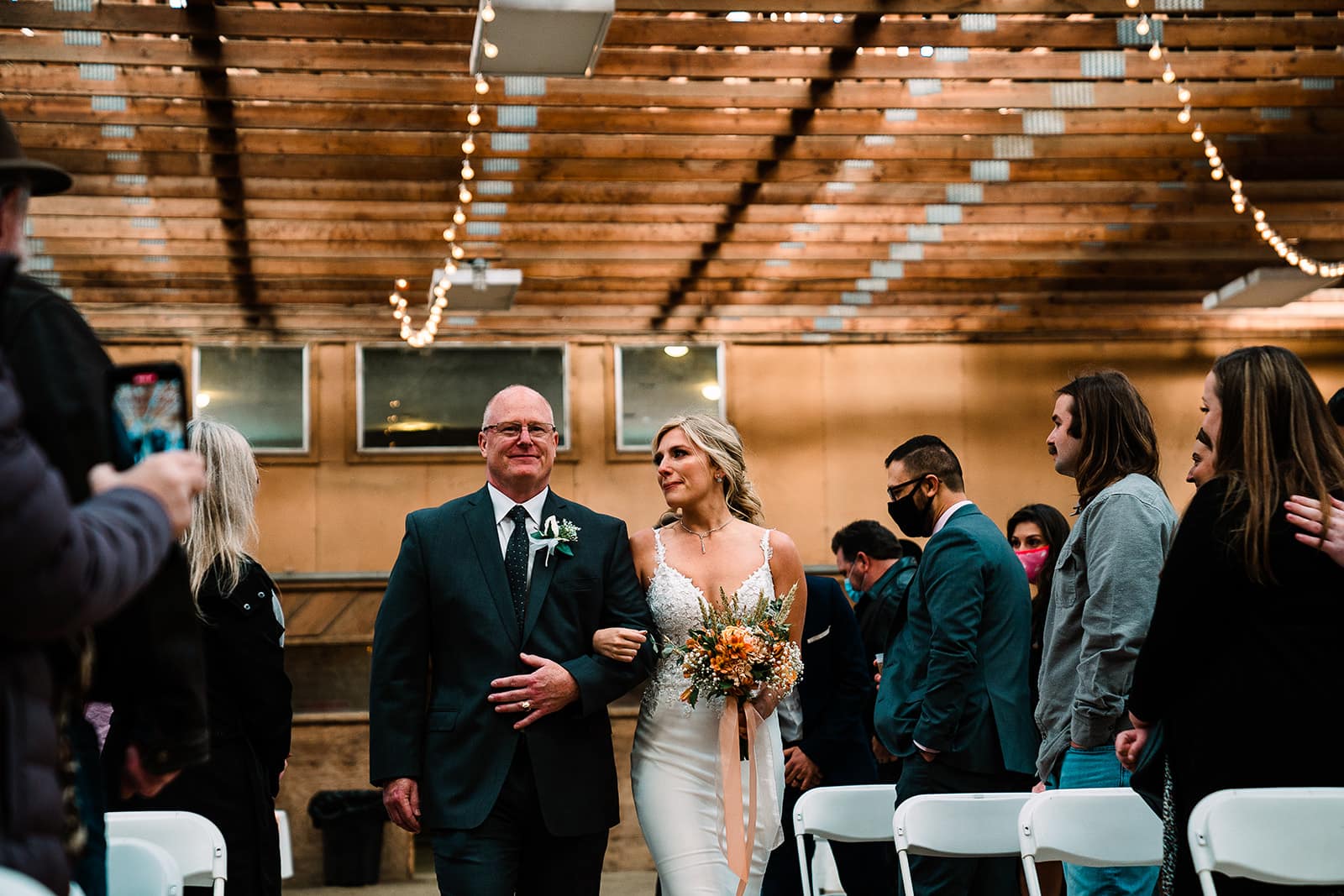 Bride walking down the aisle with her father during a wedding ceremony, surrounded by guests in a decorated venue with string lights and a rustic atmosphere. The bride holds a bouquet and wears an elegant wedding dress, while her father is dressed in a suit.