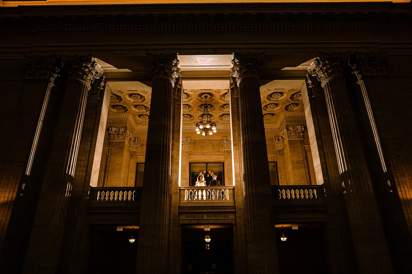 Investment 2 Couple standing on a balcony in a grand building, illuminated by soft lighting, surrounded by tall columns and ornate architectural details.