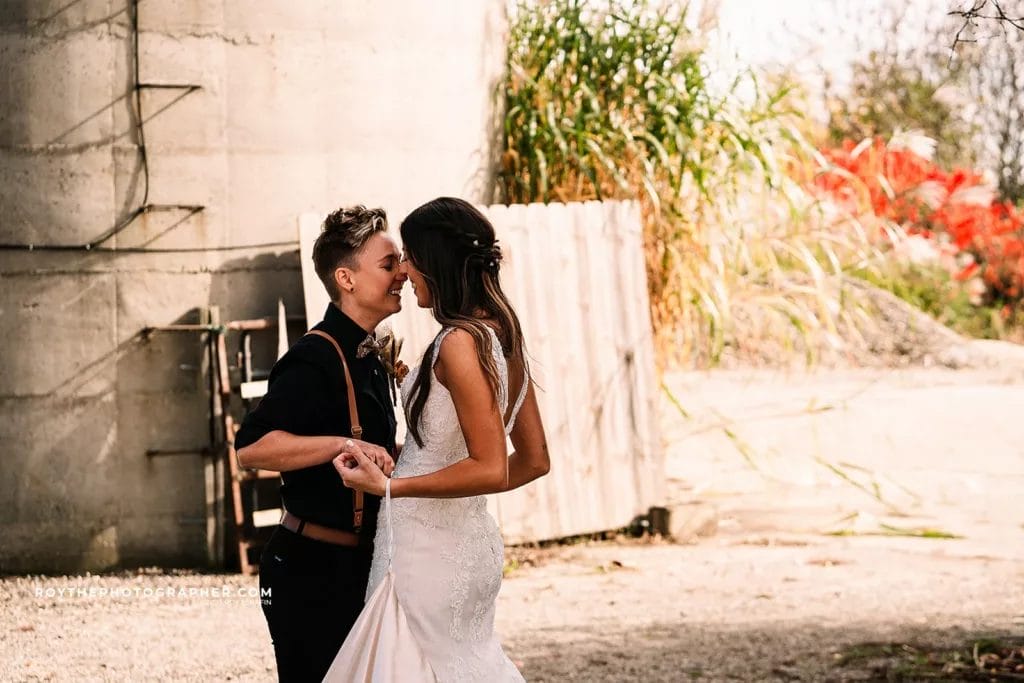 Same-sex couple sharing a romantic moment outdoors, embracing and smiling at each other, with a rustic background featuring greenery and a wooden fence. The person on the left is wearing a black shirt with suspenders, while the person on the right is dressed in a white wedding gown.
