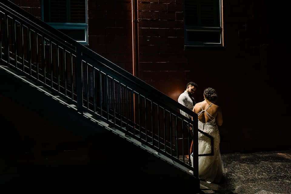 Wedding Experience 9 Couple sharing an intimate moment on a staircase at night, illuminated by soft lighting, showcasing a wedding dress and groom's attire against a dark brick wall.