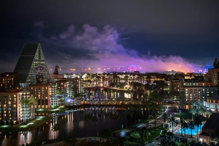 Orlando skyline at night featuring the Swan and Dolphin Resort, with illuminated buildings, palm trees, and a view of EPCOT's Spaceship Earth in the background. The scene reflects a vibrant atmosphere with colorful lights and water reflections.