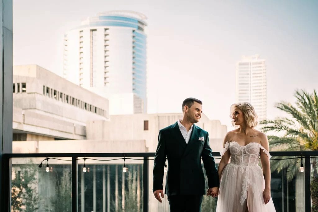 A couple stands on the balcony in wedding attire and holding hands with the backdrop of Downtown Orlando.