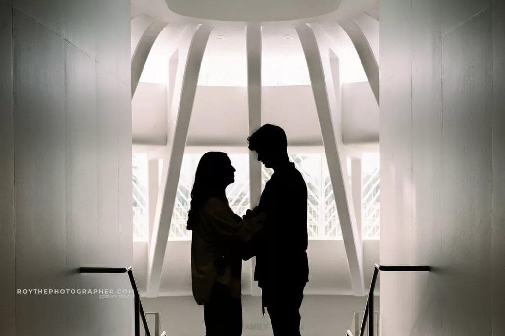Silhouette of a couple embracing in a modern architectural space with bright light filtering through a unique ceiling design, creating a romantic atmosphere.