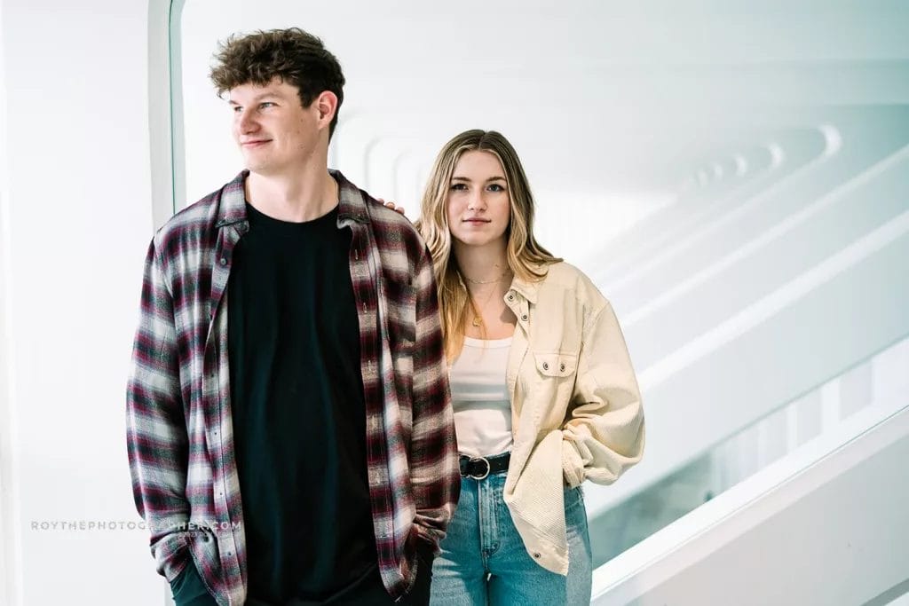 Two young adults pose for a portrait in a modern interior space with white walls and geometric lines. The male, wearing a plaid shirt over a black t-shirt, stands beside a female dressed in a beige oversized shirt and jeans. The female has long, wavy hair and a subtle smile, while the male looks off to the side.