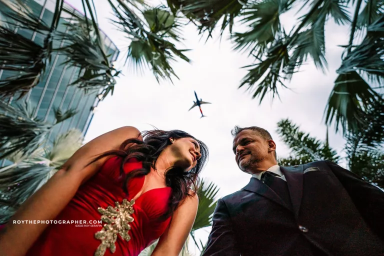 A couple in formal attire look up towards the sky framed by palm leaves at the Lake Nona Wave Hotel, with an airplane soaring directly above them.