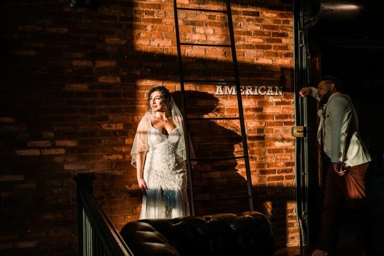 Bride in a wedding dress with intricate detailing stands against a brick wall, illuminated by natural light, while the groom leans casually against a doorframe. The setting features a rustic ambiance with a ladder and a sign that reads "The American Room."