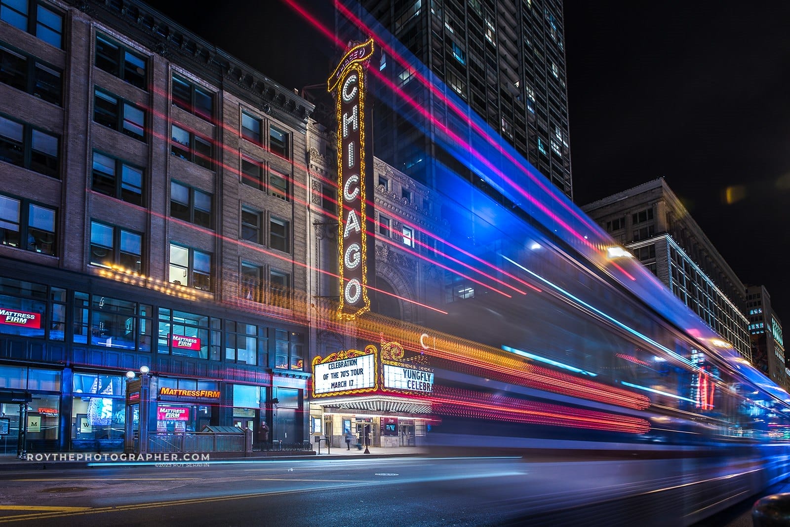 About 8 Nighttime view of the Chicago Theatre illuminated with bright marquee lights, showcasing a performance advertisement. Traffic trails create a dynamic effect, emphasizing the city's vibrant nightlife. Nearby buildings, including a Mattress Firm store, add to the urban landscape.