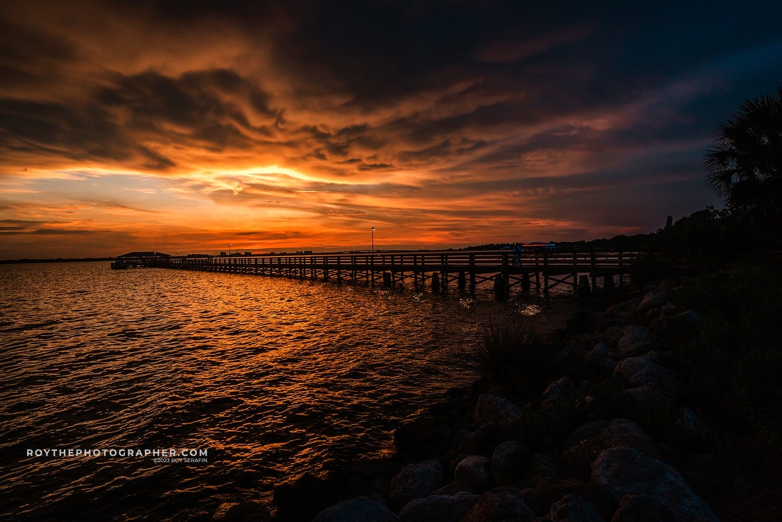 About 7 Sunset over a tranquil lake with a wooden pier extending into the water, surrounded by rocky shoreline and silhouetted clouds. The scene features vibrant orange and purple hues reflecting on the water's surface.