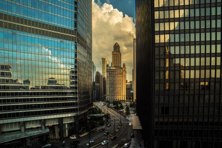 City skyline view featuring modern glass buildings and a historic tower under a cloudy sky. The image captures a bustling street with vehicles and greenery, highlighting urban architecture and city life.