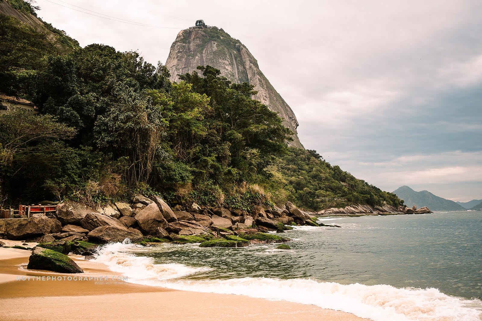 About 5 Coastal view featuring a sandy beach, rocky shoreline, and lush greenery with a prominent mountain in the background. The mountain is topped with a cable car station, under a cloudy sky.