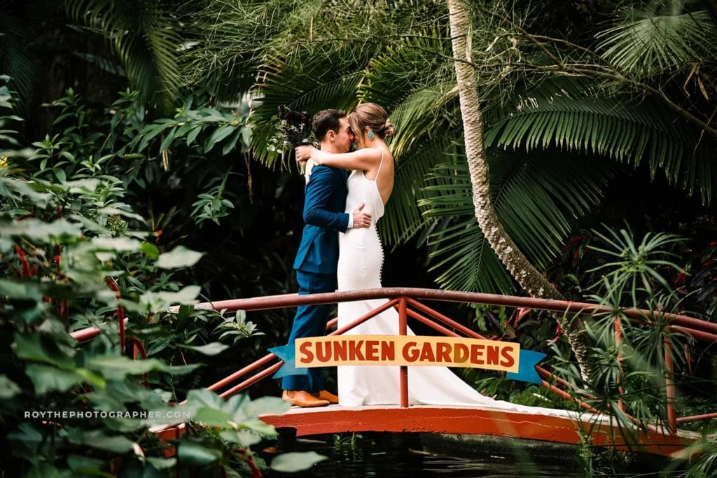A couple embraces on a red bridge at Sunken Gardens, surrounded by lush tropical greenery, with the "Sunken Gardens" sign prominently displayed in the foreground.