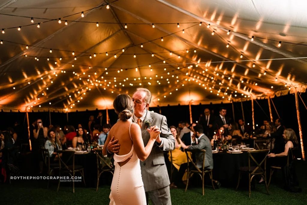 The bride shares a dance with her father at her Sunken Gardens wedding, with guests looking on in a warmly lit tented reception area.