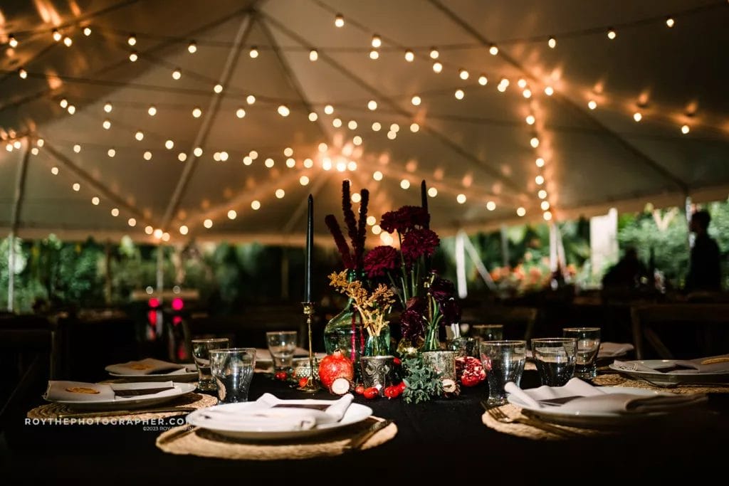 A beautifully arranged wedding reception table under a tent at Sunken Gardens, with elegant string lights above, and a centerpiece featuring deep red flowers and candles.