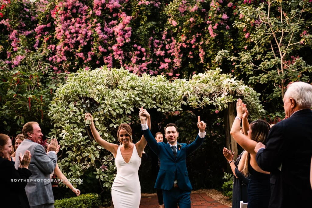 The bride and groom joyously walk down the aisle after their wedding ceremony at Sunken Gardens, with guests clapping and vibrant pink blooms in the background.