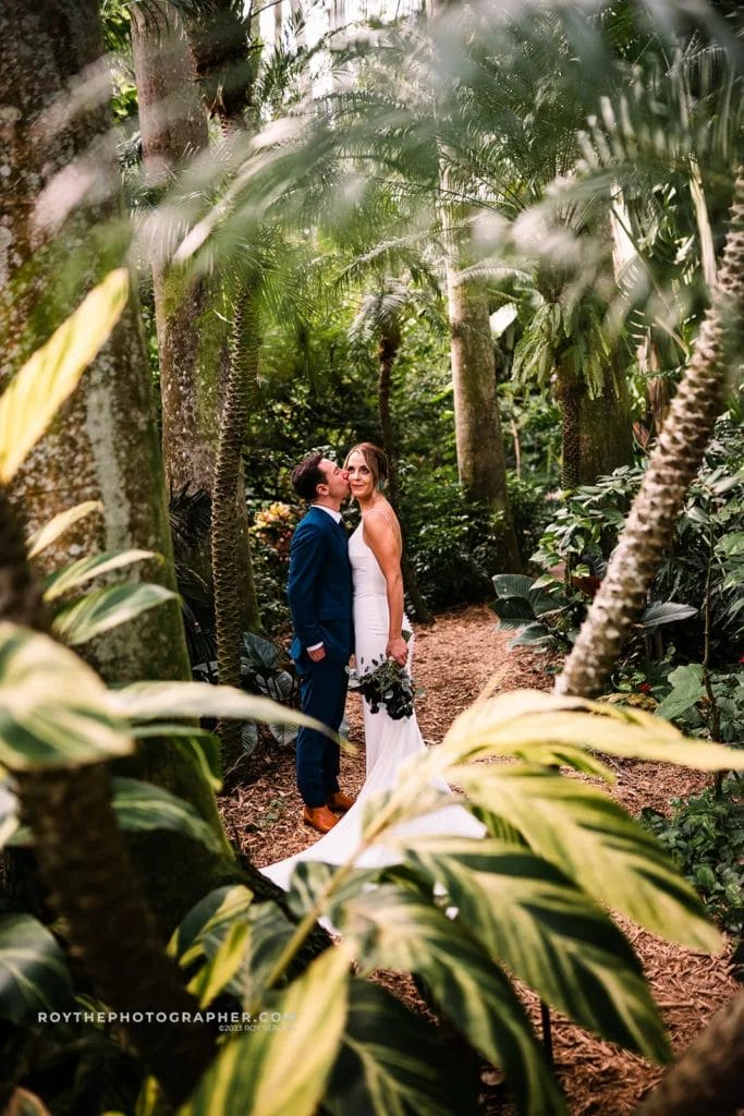 A loving couple shares a quiet moment on a secluded path at Sunken Gardens, enveloped by towering palms and tropical plants, celebrating their wedding day.