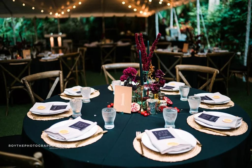 A Sunken Gardens wedding reception table beautifully set for guests, with elegant menu cards and golden accents against a backdrop of twinkling lights and lush greenery.