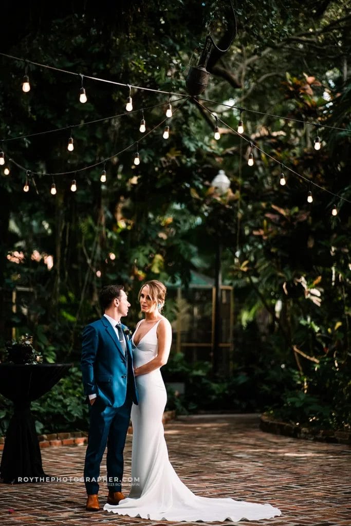 The couple stands close together at Sunken Gardens, exchanging a loving gaze, framed by the evening's soft light filtering through the canopy and delicate string lights above.