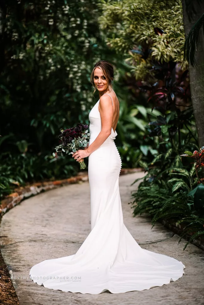 A bride in a sleek white gown holds a dark bouquet, standing on a winding path within the verdant foliage of Sunken Gardens, looking over her shoulder with a serene expression.