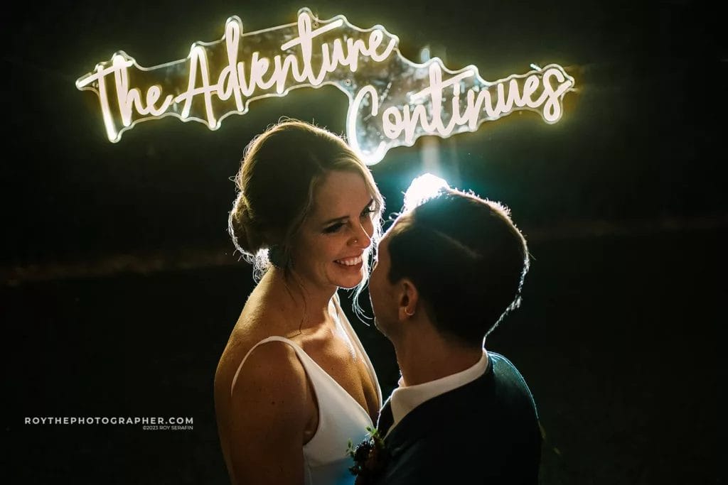 A bride and groom stand under a bright neon sign that reads "The Adventure Continues," symbolizing the start of their new journey together, post-Sunken Gardens wedding celebration.