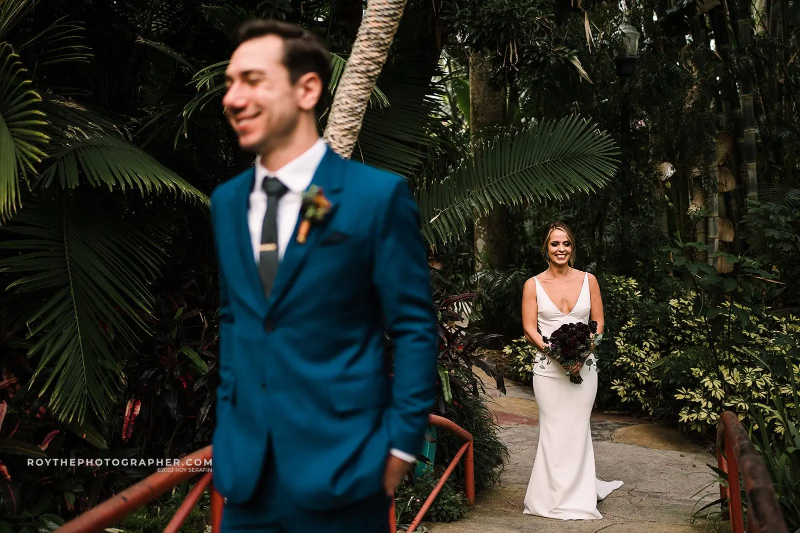 A bride beams with happiness as she looks at her groom, who stands in the foreground, out of focus, at their Sunken Gardens wedding, with lush greenery surrounding them.