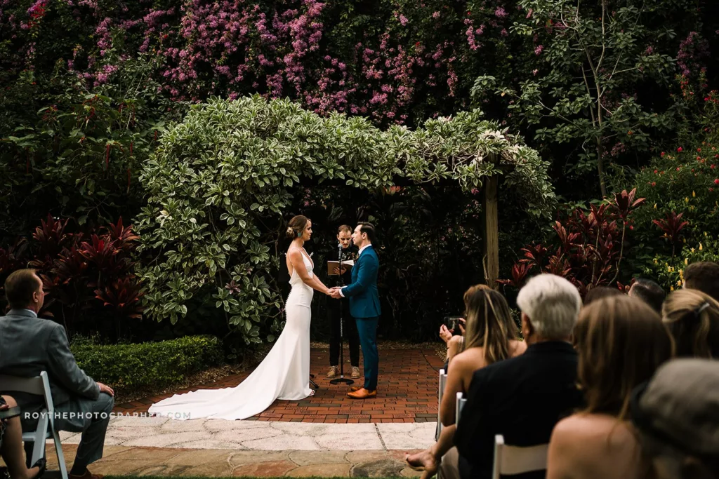 A close-up of the bride and groom holding hands during their Sunken Gardens wedding ceremony, with the focus on their intimate connection and the bride's elegant gown.