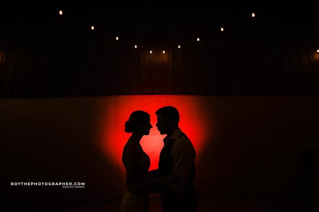 Silhouette of a couple in formal attire embracing under warm ambient lighting, with a red backdrop and string lights overhead, creating a romantic atmosphere.