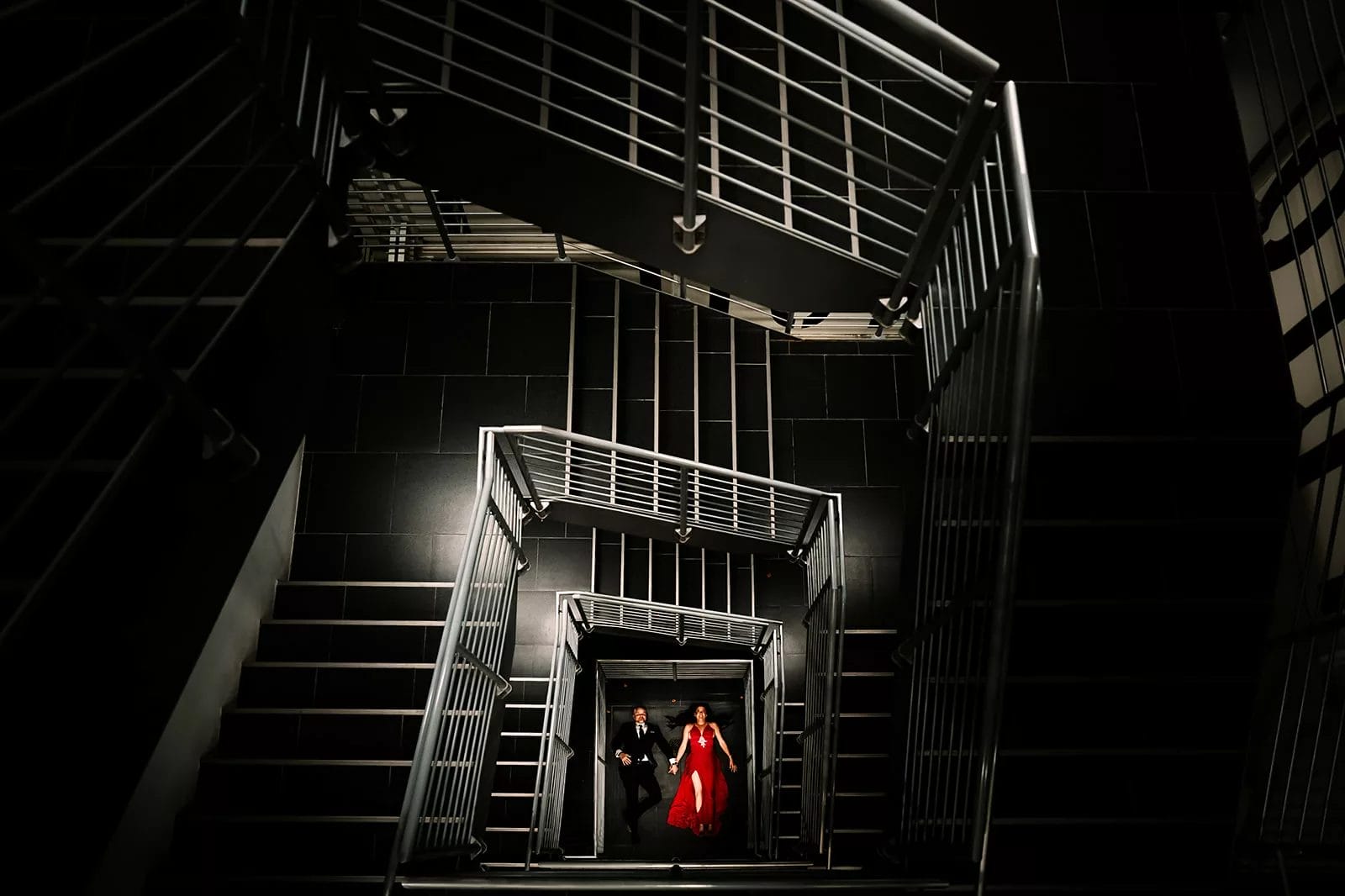 Couple walking down a modern staircase, featuring sleek metal railings and dark tiled floors, with one partner wearing a red gown and the other in formal attire.