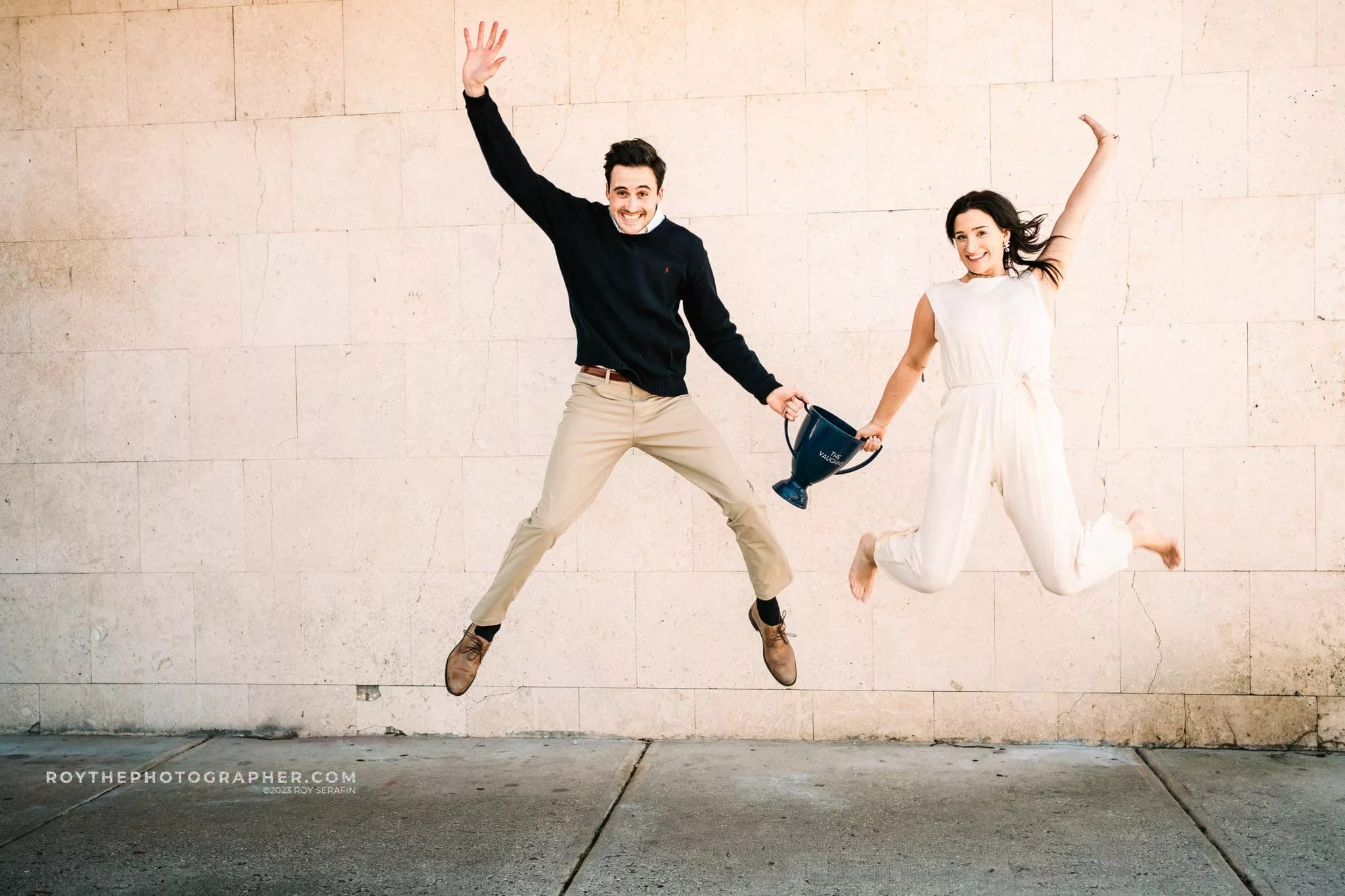 A man and woman joyfully jumping in front of a textured stone wall, with the man holding a trophy. The woman is wearing a white jumpsuit, while the man is dressed in a black sweater and beige pants. The image captures a moment of celebration and excitement.