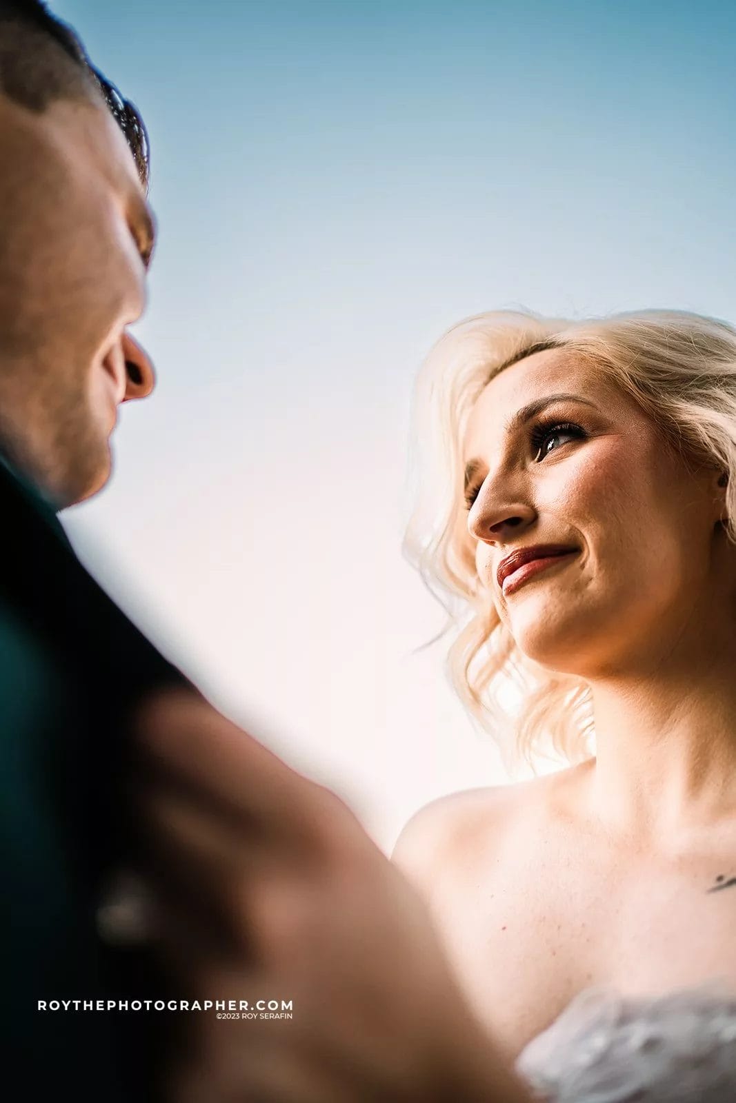 Couple gazing at each other in an intimate moment, with the bride wearing a strapless wedding gown and the groom in a dark suit, against a soft blue background.