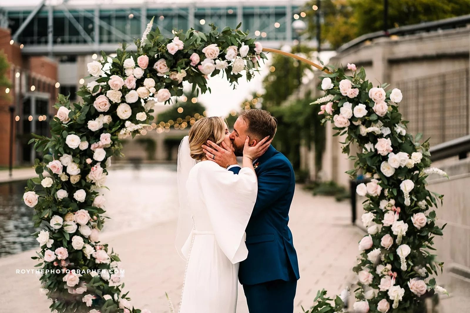 Couple sharing a kiss under a floral arch during their wedding ceremony by a river, surrounded by greenery and soft lighting.