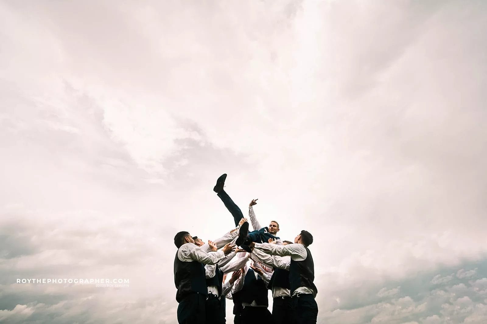 Groom being lifted by groomsmen in formal attire against a cloudy sky, capturing a joyful wedding celebration moment.
