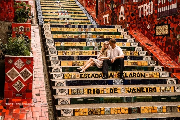 Couple sitting on colorful tiled steps in Rio de Janeiro's Selarón Steps, surrounded by vibrant mosaics and greenery.