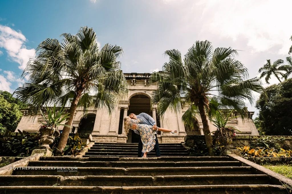 A couple in brazil in front of a mansion where the man is dipping and kissing the woman. 