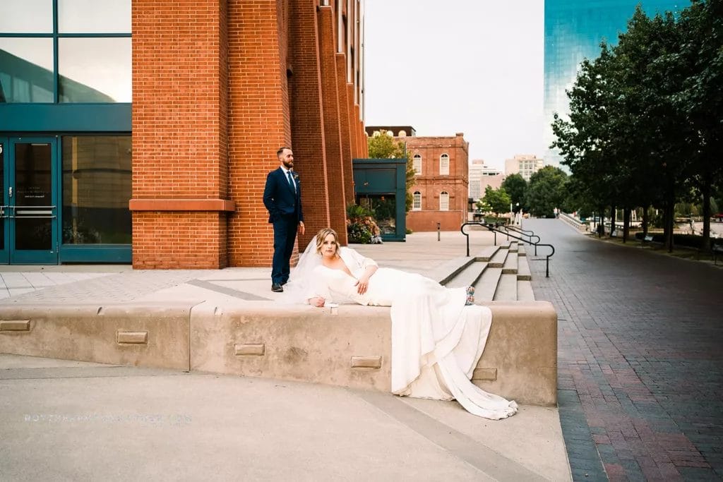 Bride laying on the ground looking at the camara while husband looks off to the park nearby. 