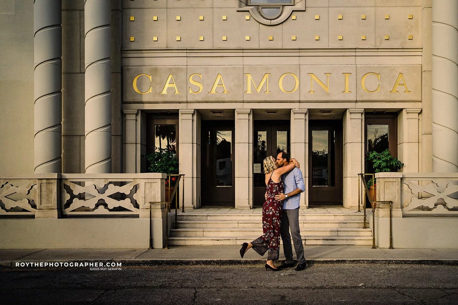 Couple embracing in front of Casa Monica, a historic hotel with ornate architecture, in St. Augustine, Florida. The image captures a romantic moment with the couple standing on a sidewalk, showcasing the building's elegant entrance and decorative columns.