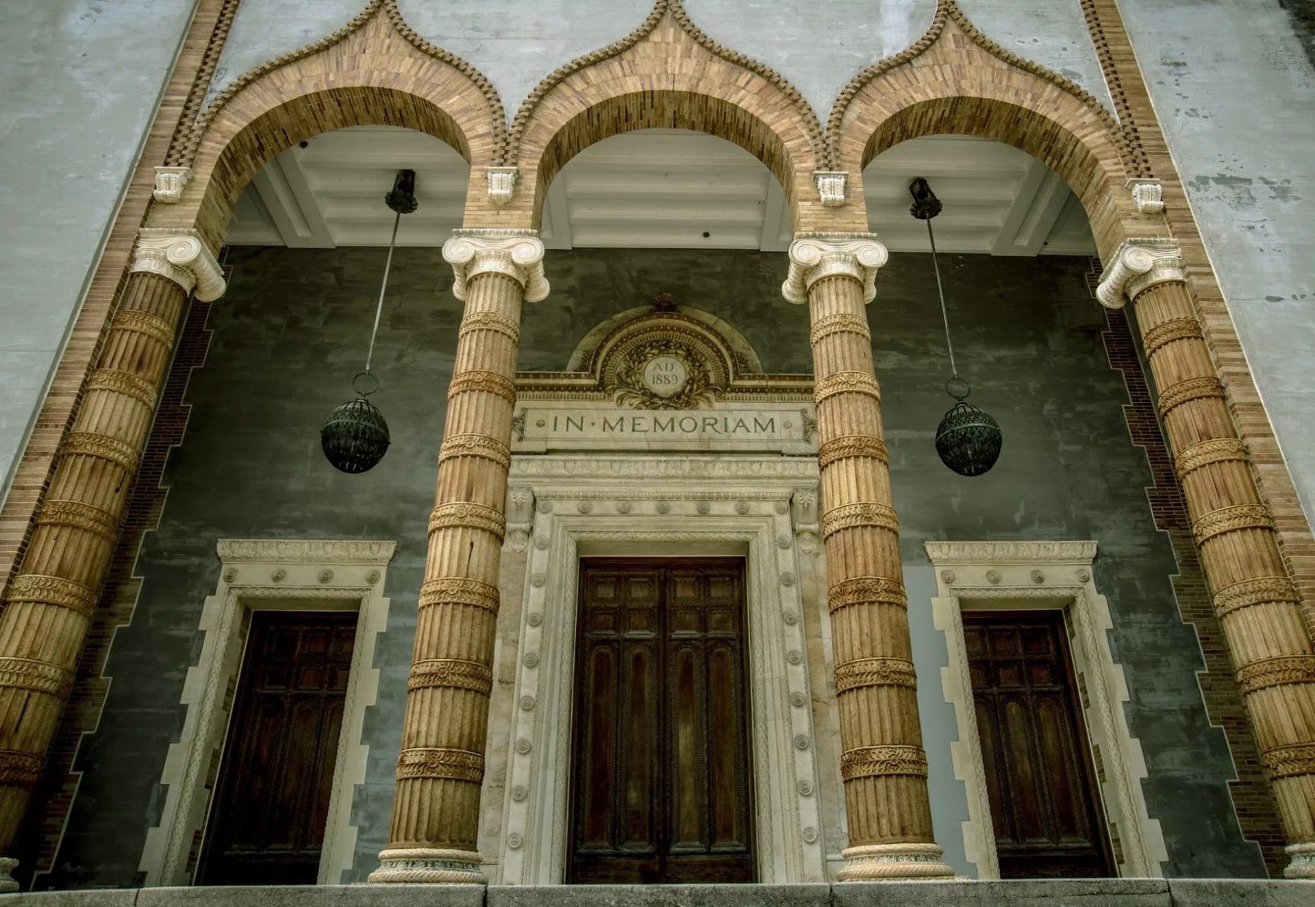 Historic building entrance featuring three ornate arches supported by intricately designed columns, with a central door marked "IN MEMORIAM" and the year "1889." Two wooden doors flank the entrance, complemented by hanging lanterns.
