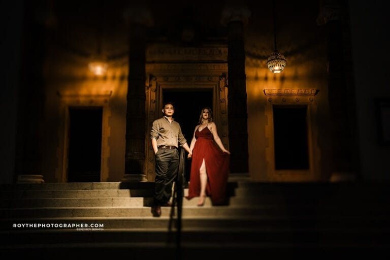Couple posing on steps in front of an ornate building at night, with warm lighting highlighting their outfits; the woman wears a red dress and the man is in a button-up shirt and pants, both holding hands.