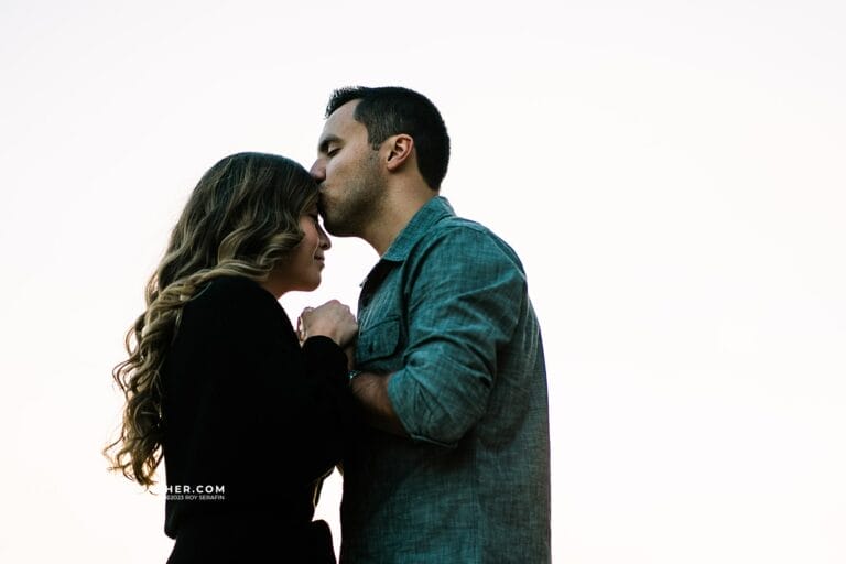 Couple sharing a tender moment, with the man kissing the woman's forehead as they hold hands, set against a soft, neutral background.