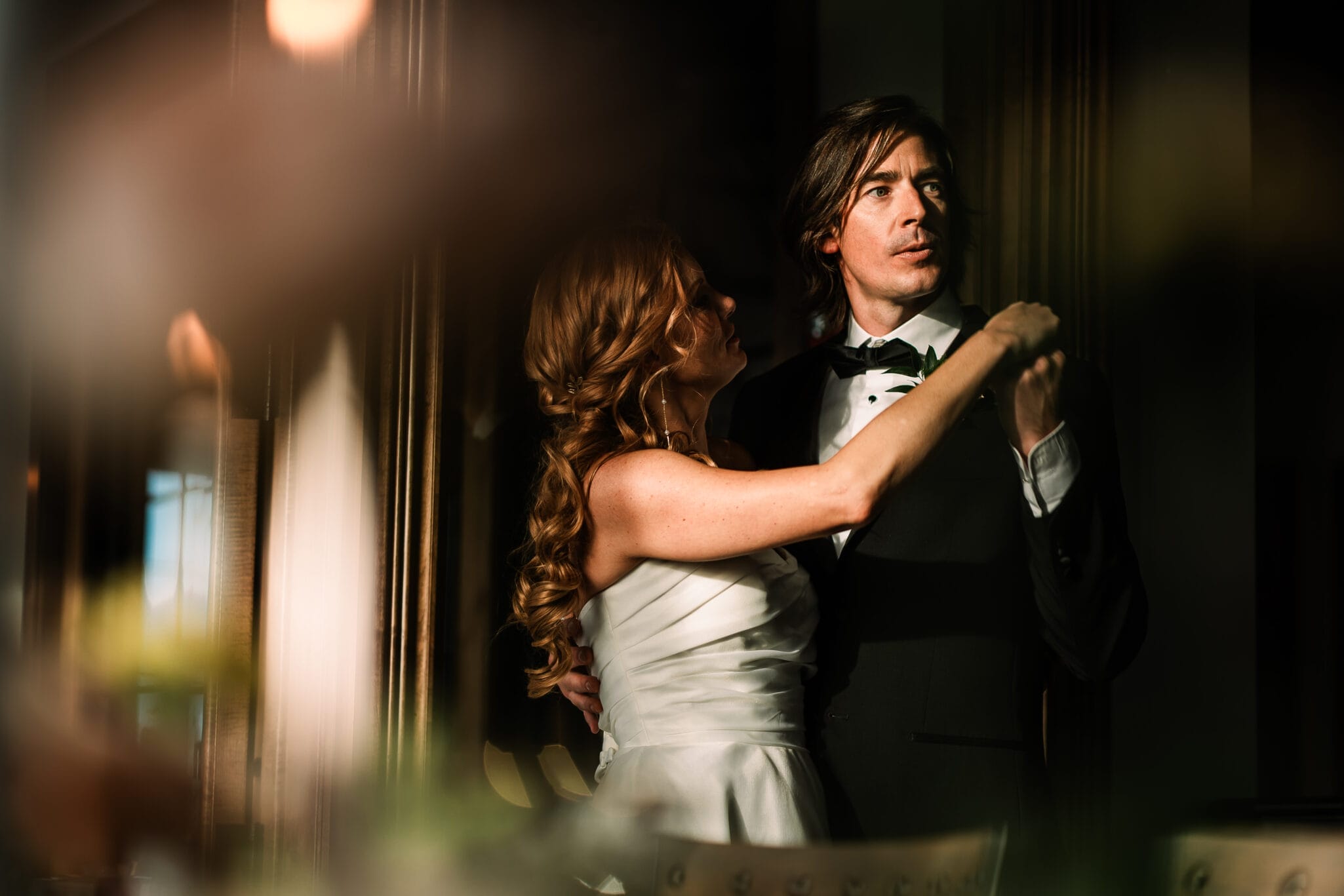 Couple in formal attire sharing an intimate moment, with the woman in a white wedding dress and the man in a black tuxedo, set against a softly blurred background.