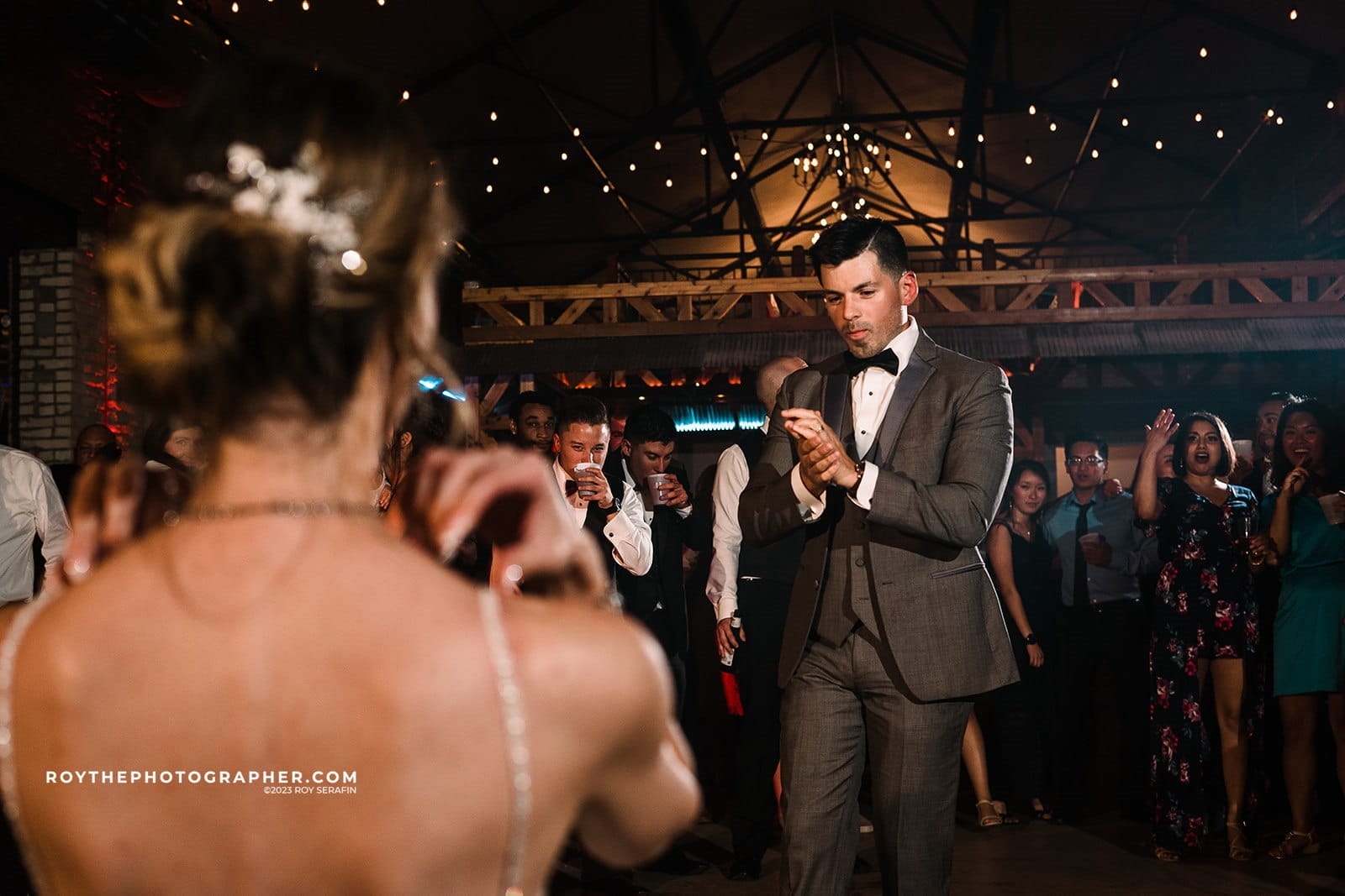 Wedding reception dance floor with a man in a tuxedo and a woman in a sparkling dress. The background features guests celebrating, clapping, and enjoying drinks under string lights.