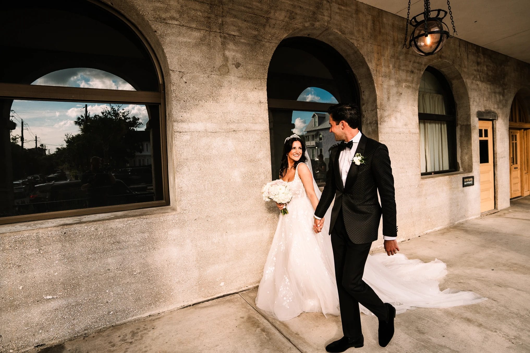 Bride and groom walk hand in hand outside a building, with the bride holding a bouquet of white flowers. The groom wears a black tuxedo while the bride is dressed in an elegant white wedding gown. The scene captures a joyful moment in an urban setting, with reflections of trees and clouds visible in nearby windows.