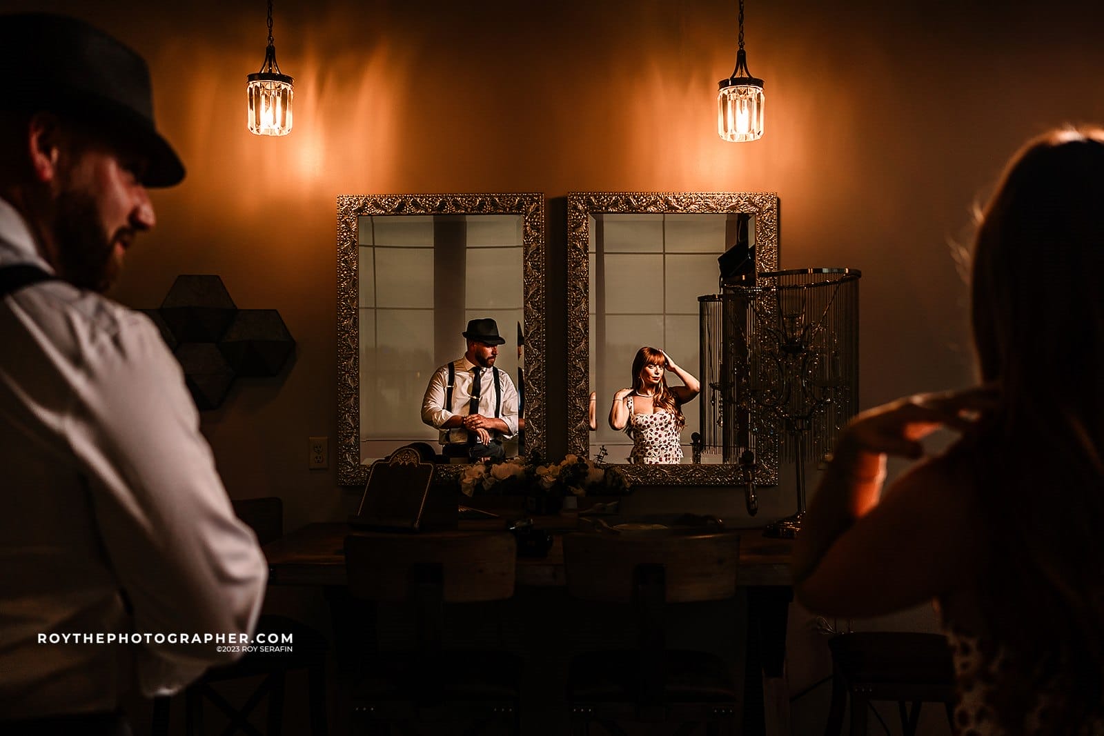 Phoenix Wedding Photographer 39 Couple preparing for an event in a stylish room with mirrors and vintage lighting. The woman adjusts her hair while the man stands by, both dressed elegantly. The setting features a wooden table and decorative elements, creating a warm atmosphere.