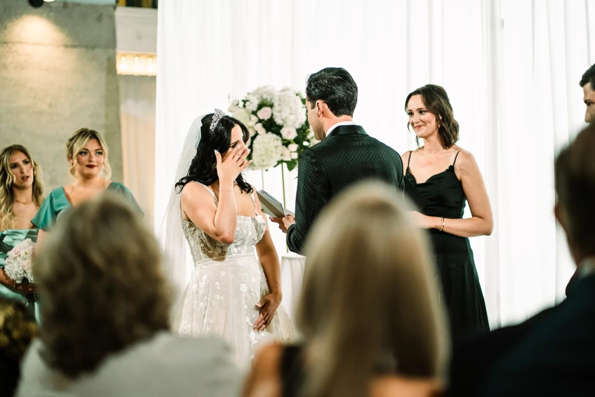 Bride and groom exchanging vows during a wedding ceremony, with emotional expressions. The bride, wearing a lace gown and a veil, shows a heartfelt reaction, while the groom stands with a wedding ring. Bridesmaids in elegant dresses and floral arrangements are visible in the background.