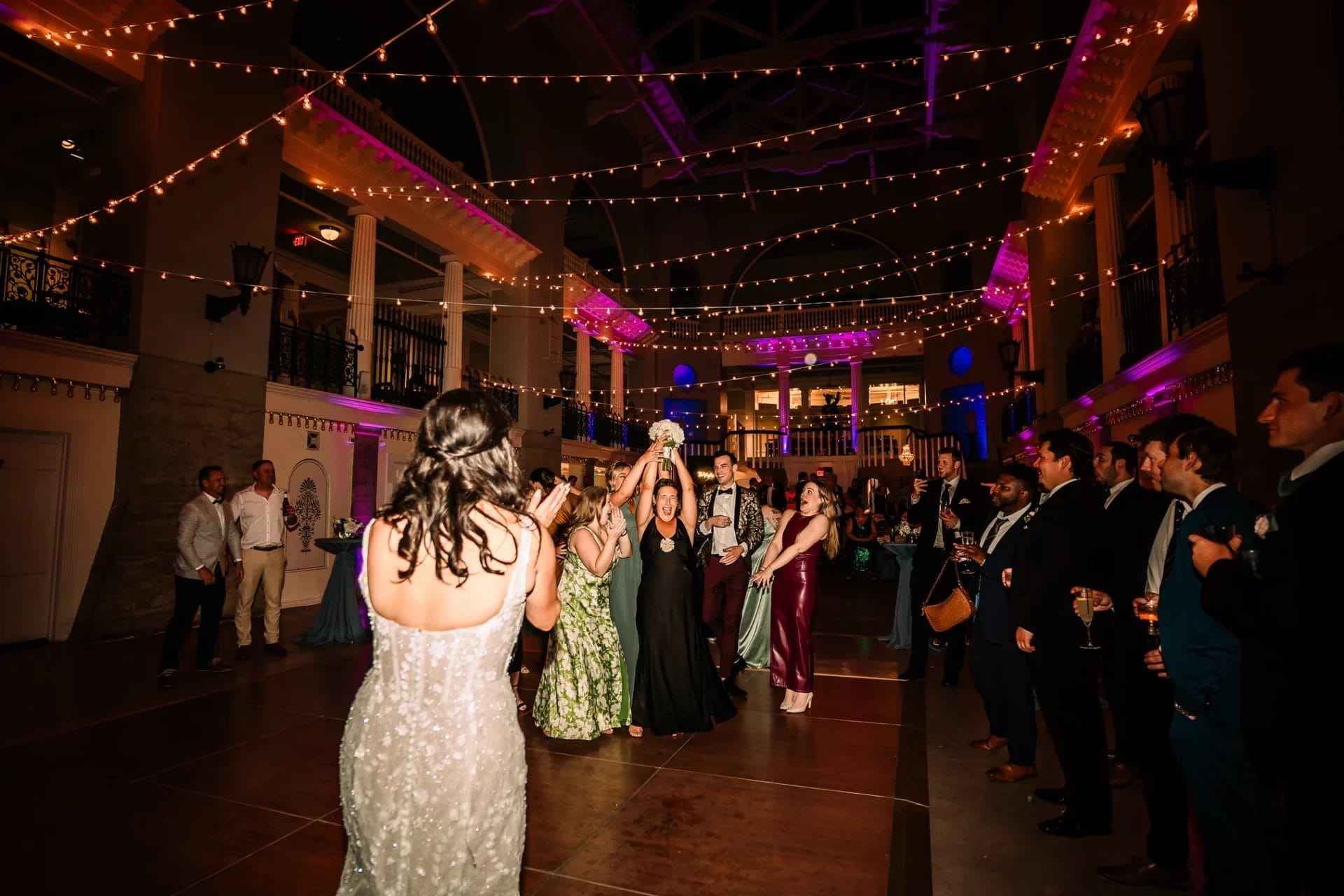 Wedding reception scene with guests dancing and celebrating under string lights. A woman in a black dress is about to throw a bouquet, surrounded by excited attendees in formal attire. The venue is elegantly decorated with ambient lighting and features high ceilings.