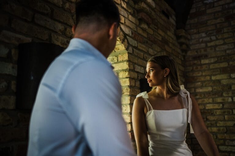 A woman in a white dress with unique shoulder details gazes out of a brick archway, with a man slightly blurred in the foreground, set in an intimate, softly lit interior space.