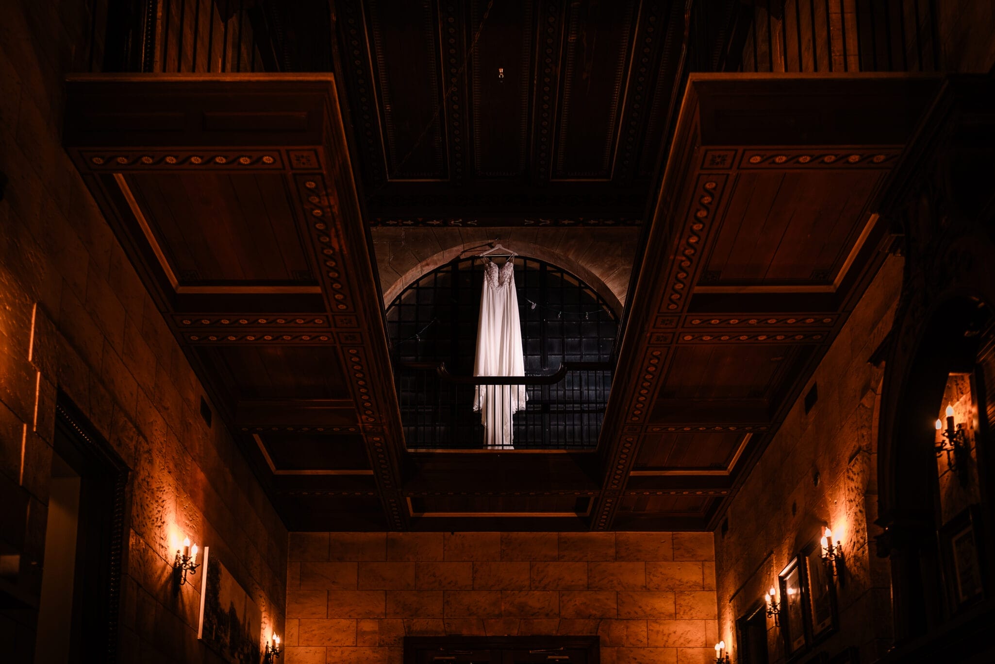Bridal dress hanging on a wooden beam in an elegant venue with warm lighting and ornate architecture, showcasing a romantic wedding atmosphere.
