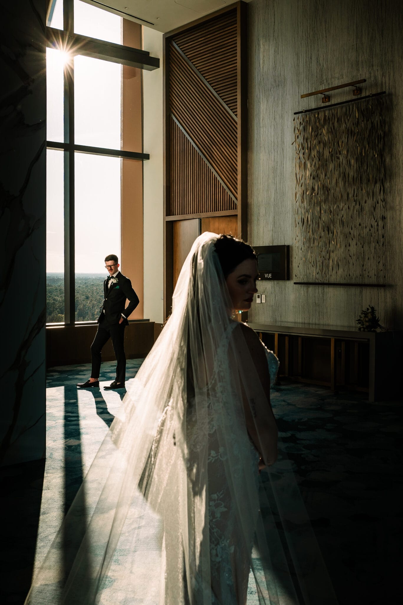 Bride in a wedding dress with a veil stands in a sunlit modern venue, while the groom, dressed in a tuxedo, poses in the background. The image highlights elegant decor and soft lighting.