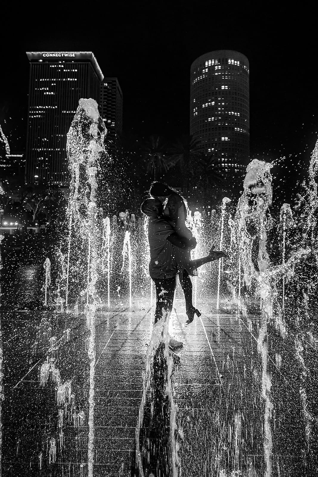 Couple embracing in a fountain at night, surrounded by illuminated water jets and city skyline, captured in black and white.