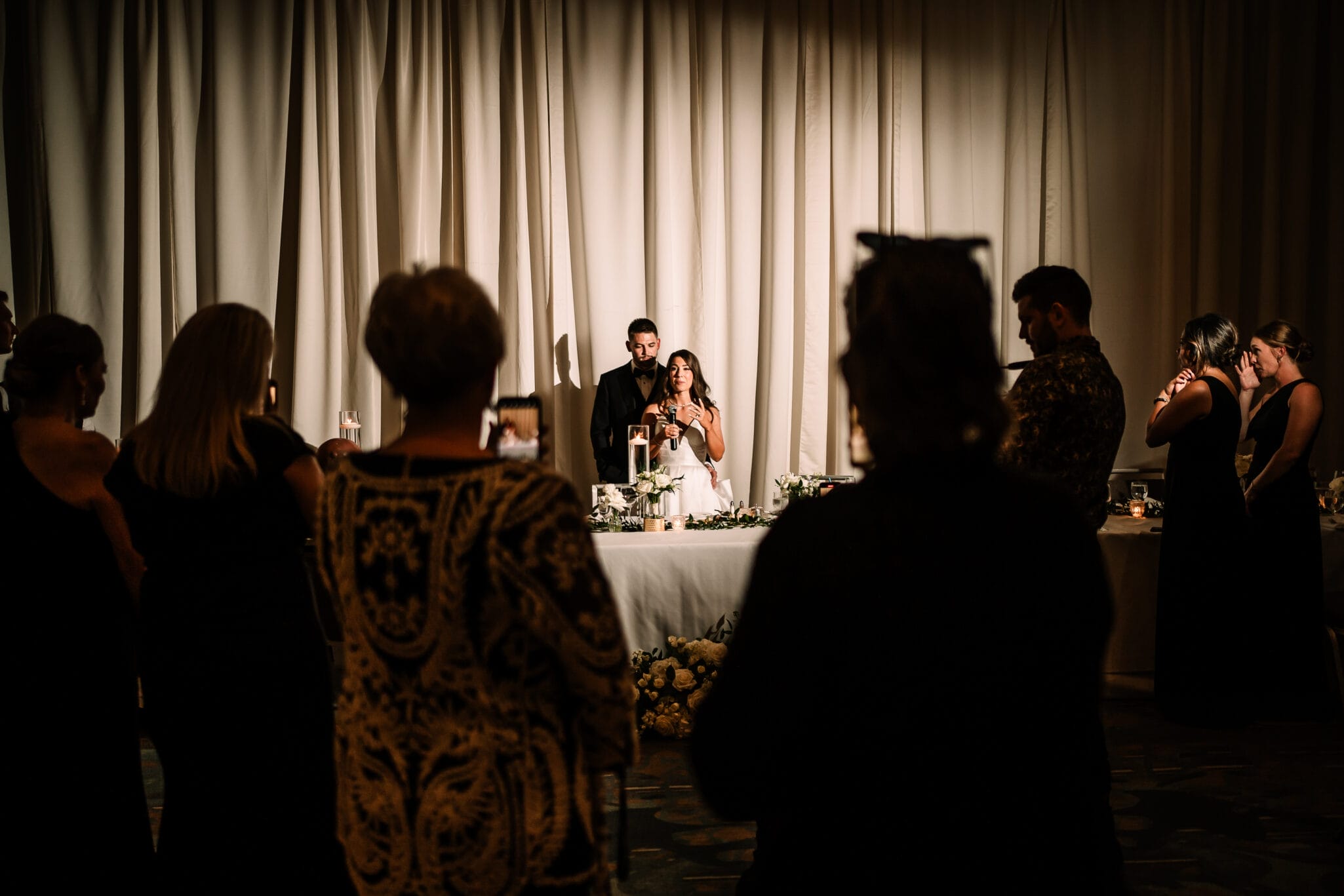 A couple stands at a wedding reception, giving a toast while guests in elegant attire listen and take photos. The setting features a decorated table with floral arrangements and candles, illuminated against a backdrop of soft drapes.